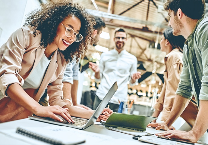 Eine Gruppe junger Berufstätiger sitzt in einem modernen Büro um einen Tisch herum, lächelt und diskutiert über Ideen. Eine Frau tippt auf einem Laptop, während die anderen sich unterhalten und so eine kooperative und energiegeladene Atmosphäre schaffen.
