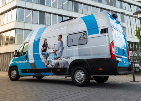 Ein blau-weißer Kleintransporter mit der Aufschrift aktivmobil und IKK Südwest steht vor einem modernen Bürogebäude, auf dessen Seitenwand zwei Personen abgebildet sind, die sich unterhalten.