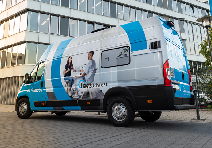 Ein blau-weißer Kleintransporter mit der Aufschrift aktivmobil und IKK Südwest steht vor einem modernen Bürogebäude, auf dessen Seitenwand zwei Personen abgebildet sind, die sich unterhalten.