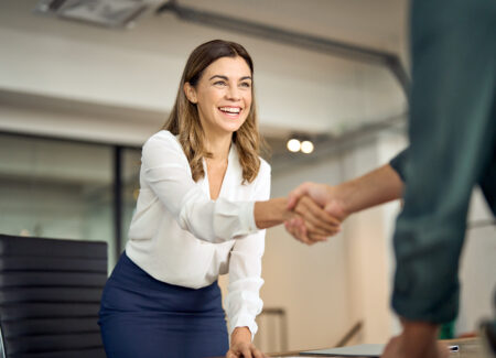 Eine lächelnde Frau in Geschäftskleidung beugt sich über einen Schreibtisch, um einer anderen Person in einem Büro die Hand zu geben.