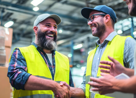 Drei Männer mit gelben Sicherheitswesten und Mützen lächeln und schütteln sich die Hände in einem Lagerhaus, das von Kisten und Industrieregalen umgeben ist und ein positives und freundliches Arbeitsumfeld zeigt.